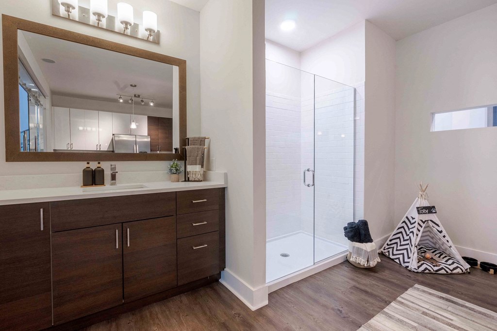 Studio Apartments in Tampa, FL - Society Westshore - Bathroom with Wood Plank Style Flooring, Large Vanity Mirror, Quart Countertop, Dark Wood-Tone Cabinetry, and Shower with Glass Door.