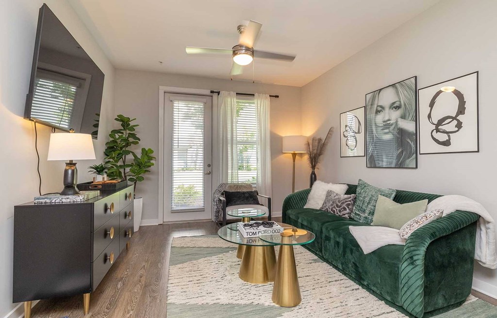 Apartments for Rent in Downtown Greenville, SC - The Aster - Living Room with Vinyl Plank Flooring, Area Rug, Green Velvet Couch, Small Round Coffee Tables, Wall Mounted TV, Ceiling Fan, and Door to Outdoor Access with Blinds and White Curtain.