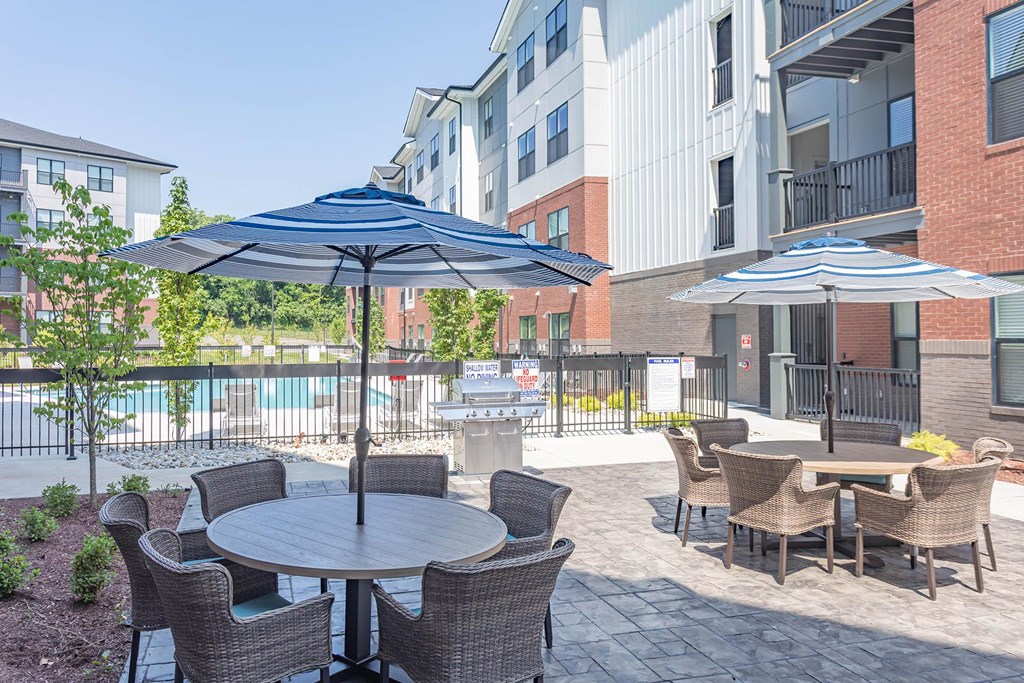 Apartments For Rent In Hermitage, TN - Outdoor Patio Area By The Pool. Patio Has Gas BBQ Grills, Tables With Umbrellas, Chairs, And Maintained Landscaping.