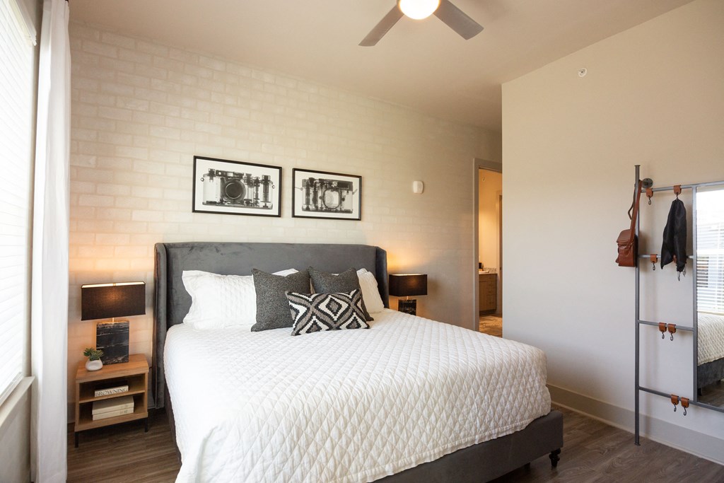 1 BR Apartments in Downtown Memphis TN - The Landing Residences - Furnsihed Bedroom wtih Hardwood Floor, Bed, Nightstands with Lamps, Standing Mirror with Storage, Ceiling Fan, White Painted Brick Accent Wall, and Attached Bathroom