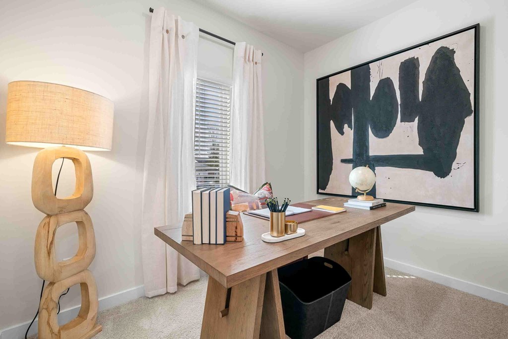 A room with a table, a lamp, a painting, and a window with curtains.