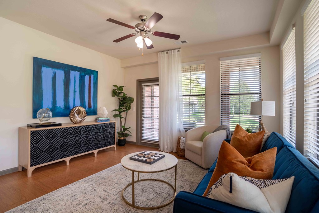 Pet Friendly Apartments in Greenville SC - Velo Verdae - Furnished Living Room with Natural Oak-Style Plank Flooring, Area Rug, Blue Sofa, White Armchair, Marble and Gold Round Coffee Table, Black and Tan TV Monitor, Standing Lamp, Large Windows with Blinds, Ceiling Fan, and House Plant in the Corner