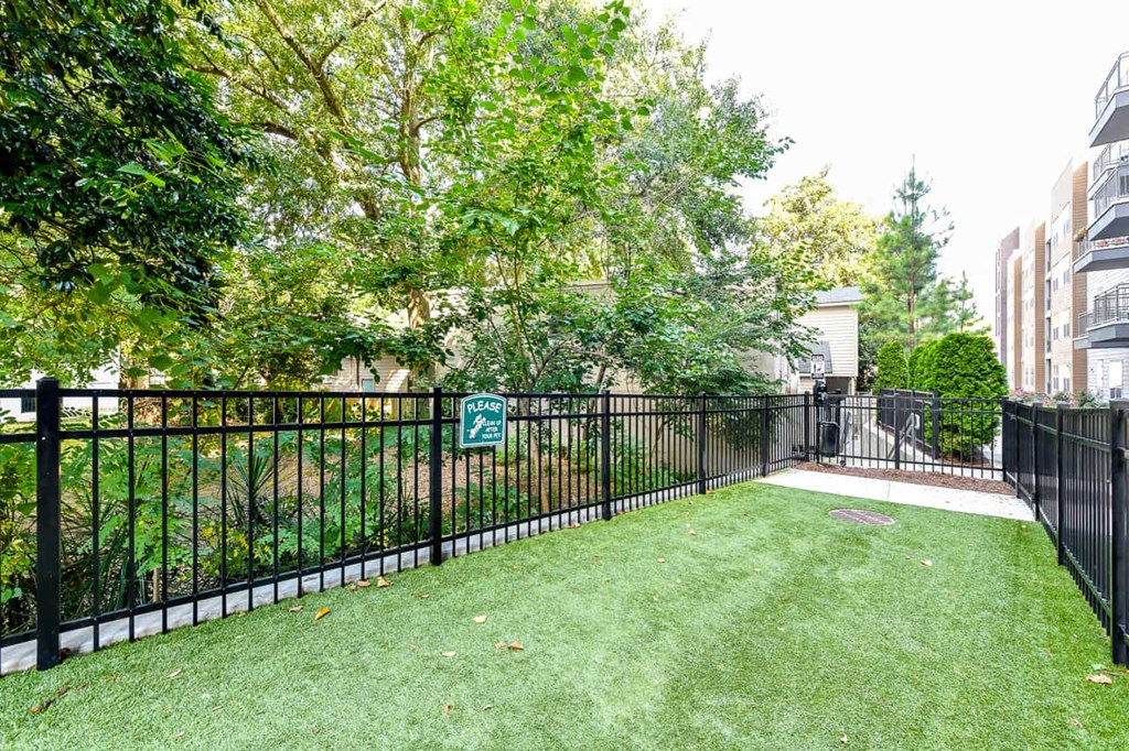 Decatur, GA Apartments Near Agnes Scott College - ARLO - Fenced Dog Park With Manicured Grass, Waste Disposal Area, And Surrounding Lush Greenery.