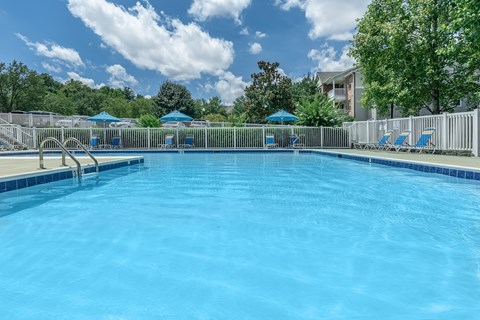 Affordable Apartments for Rent in Richmond City, VA - Overlook at Falling Creek - A Clear Outdoor Swimming Pool With Lounge Chairs, Blue Umbrellas, White Fence, and Trees.