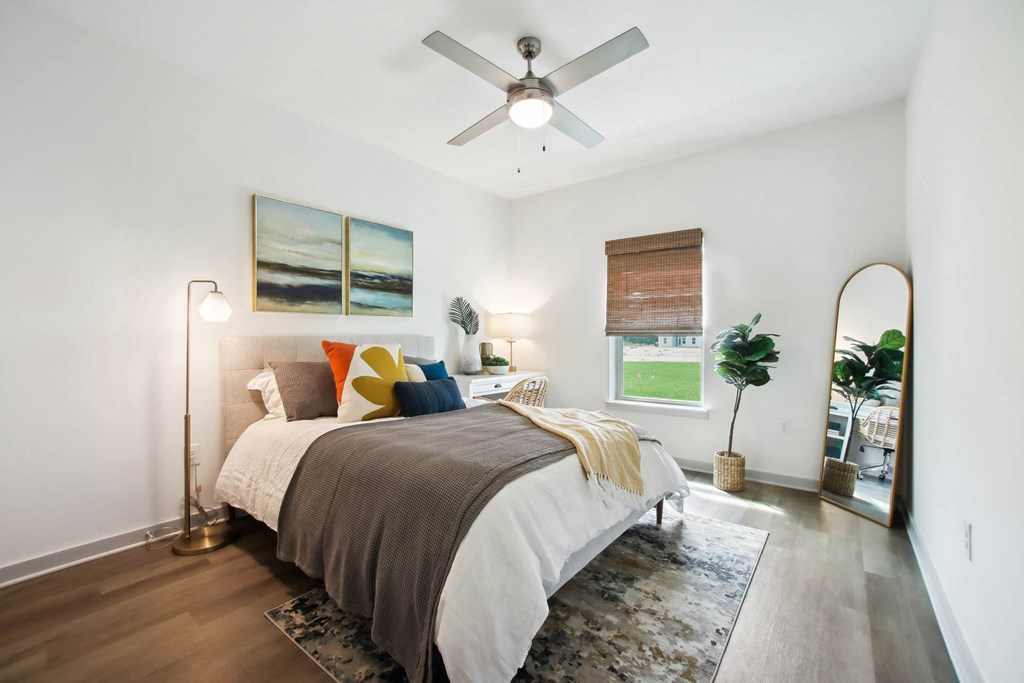 Flagler Beach, FL, Luxury Apartments with In-Unit Washer and Dryer - Preserve at Flagler Beach - Bedroom with Wood-Inspired Flooring, Area Rug, Bed, Floor Lamp, Nightstand with Lamp, Standing Mirror, Ceiling Fan, and Window.