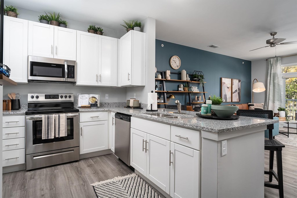 Apartments for Rent in Alexandria, KY - Elevation Apartment Homes - Kitchen with 9-Foot Ceiling, Wood-Style Flooring, White Shaker-Style Cabinetry, Stainless-Steel Frigidaire Appliances, Granite Countertops, Kitchen Tools, Decorative Plants, Eat-in Kitchen with Breakfast Bar, Bar Stool, and a View of the Living Area and Balcony Doorway.