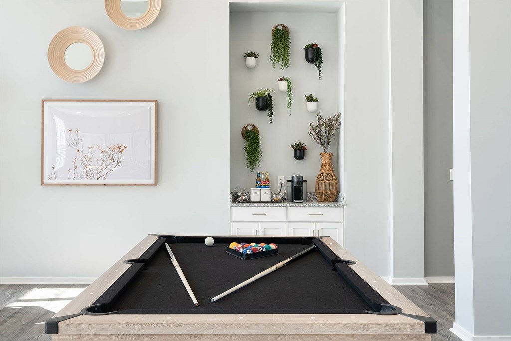 A pool table in a room with a framed picture and a shelf with plants and vases.