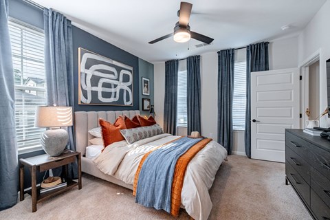 Greenville, SC, Luxury Apartments with an In-Unit Washer and Dryer - Evolve Homestead Apartment Homes - Bedroom with Large Bed, Carpet Flooring, Nightstand, Lamp, Ceiling Fan, and Dresser.