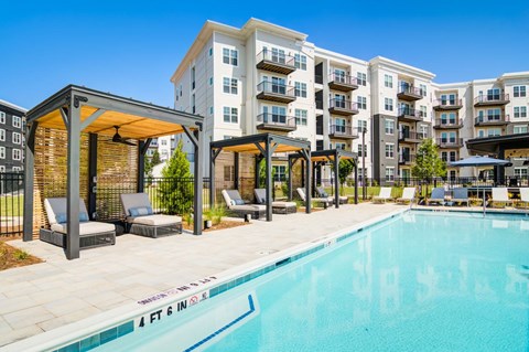 A swimming pool in front of apartment buildings with lounge chairs and a pergola.