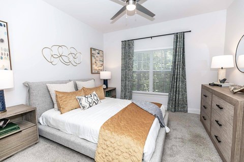 Two-Bedroom Apartments in Newnan, GA - The Jackson - Bedroom With A Gray Upholstered Bed, Gold Accent Pillows, Wood Nightstands, Ceiling Fan, Window Blinds, And Matching Dresser.