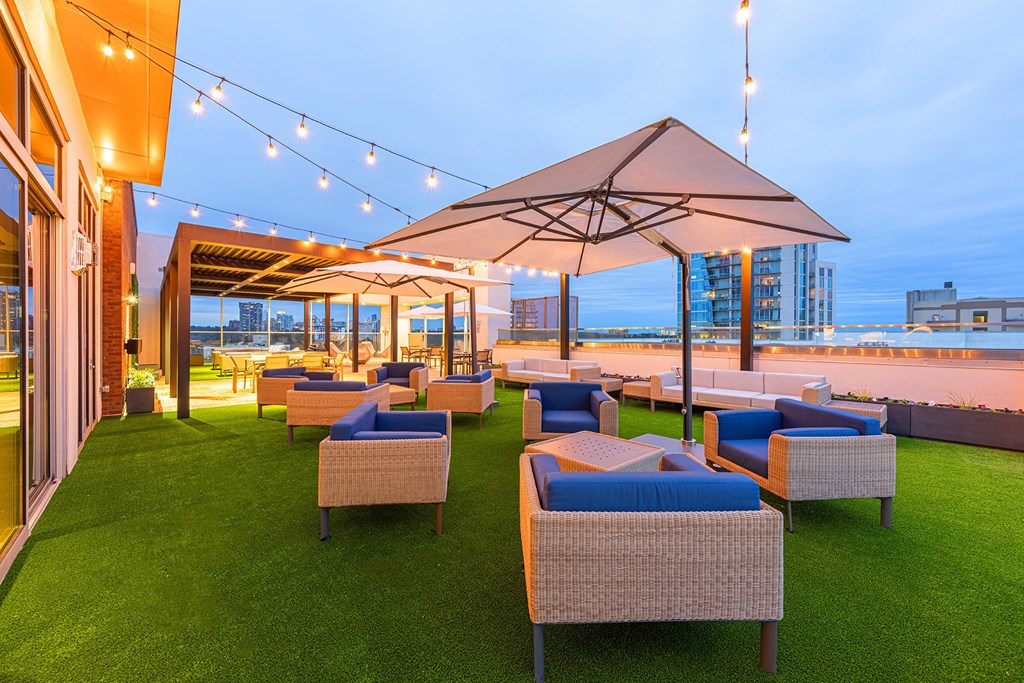 Two-Bedroom Luxury Apartments In Atlanta, GA - Venue Brookwood - Rooftop Terrace With City Skyline Views, Turf Grass, Lounge Seating, Umbrellas, Pavilion, And Cafe Lights.