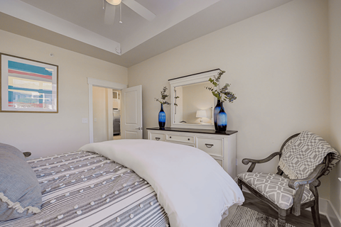1-Bedroom Luxury Apartments in Huntersville, NC - Apartments at Holly Crest - Bedroom With Patterned Bedding, White Dresser, Wall Mirror, Blue Vases, Accent Chair, And Ceiling Fan.