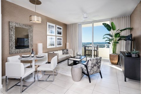 A living room with a white dining table and chairs, a sofa, a television, and a large window with a view of the ocean.