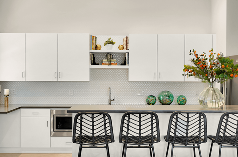 A kitchen with white cabinets and a grey backsplash with a sink and three chairs.