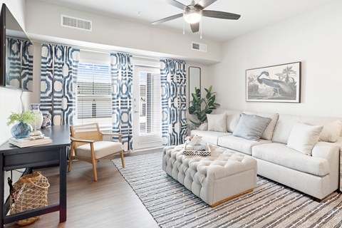 Fort Worth, TX, Senior Apartments Near Bear Creek Park - Regatta 55 - Living Room With Sofa, Armchair, Tufted Ottoman, Patterned Curtains, Ceiling Fan, and Balcony Door.