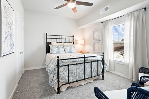 Fort Worth, TX, Senior Apartments Near Interstate 35 West - Regatta 55 - Bedroom With Soft Carpet, Ceiling Fan, Nightstand, Lamp, and Large Window With White Curtains.