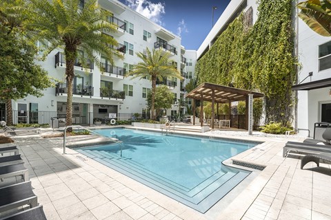 A pool surrounded by palm trees and a white building with a green wall.