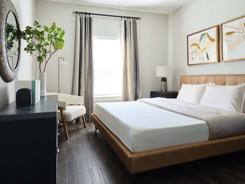 Panama City, FL Pet-Friendly Luxury Apartments - The Kelly at 23rd - Bedroom with Vinyl Wood-Inspired Plank Flooring, Queen-Size Bed, Nightstand with Lamp, Dresser, Wall-Mounted Round Mirror, Armchair, Floor Lamp, and Large Window.