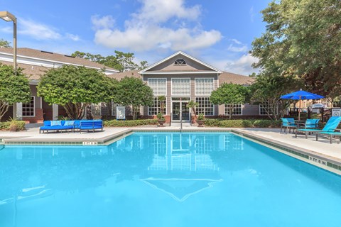 A swimming pool in front of a building with a blue umbrella and chairs.