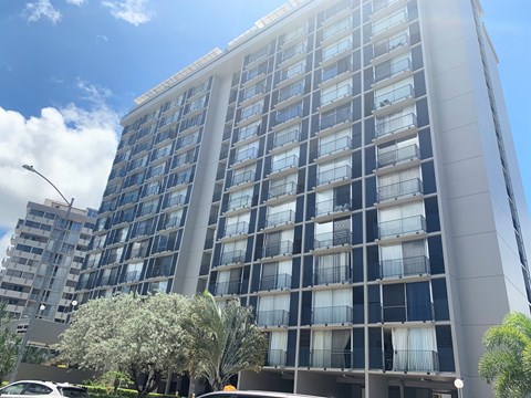 A tall building with many windows and balconies is surrounded by trees and a clear sky.