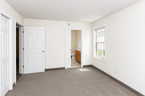 A room with a carpeted floor, a white door, a window with a white frame, and a wooden cabinet.
