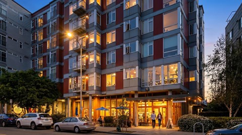 A modern apartment building with cars parked in front and people walking on the sidewalk.