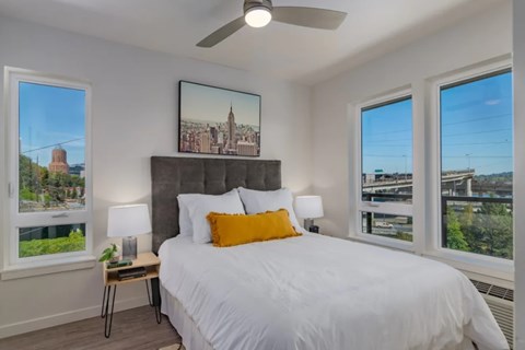 A bedroom with a large bed, a ceiling fan, and a view of a city skyline through the windows.