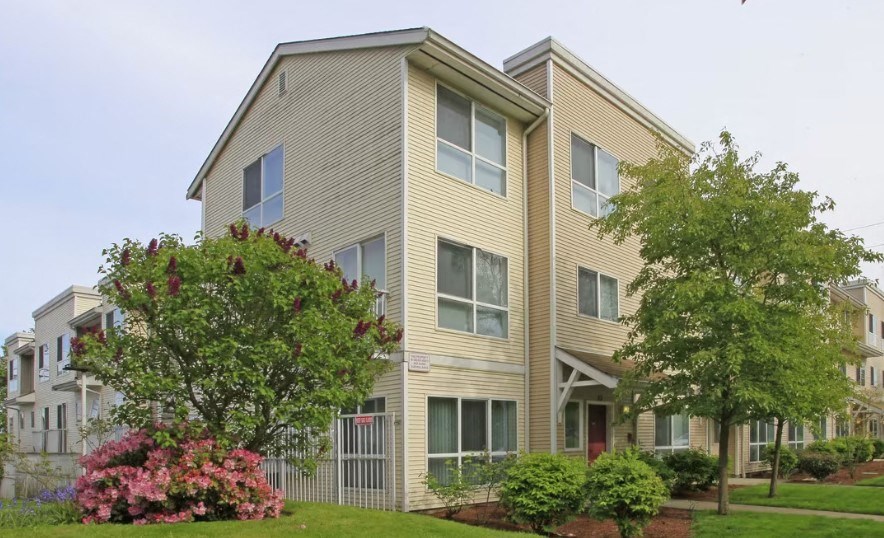 A beige two-story apartment building with a red door and a tree with pink flowers in front.