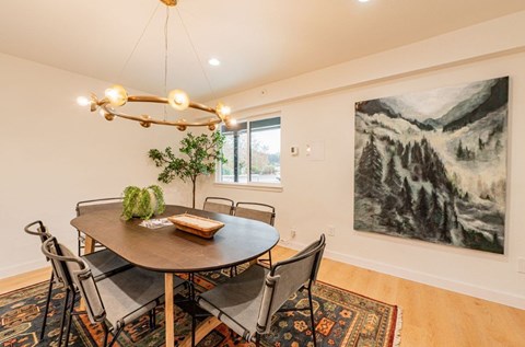 A dining room with a round table and chairs, a potted plant, and a large landscape painting on the wall.