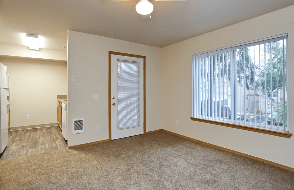 A room with a carpeted floor, a window with blinds, and a door leading to another room.