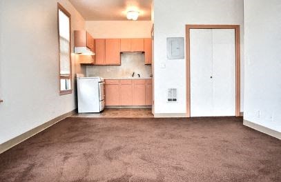 an empty living room with a refrigerator and a kitchen