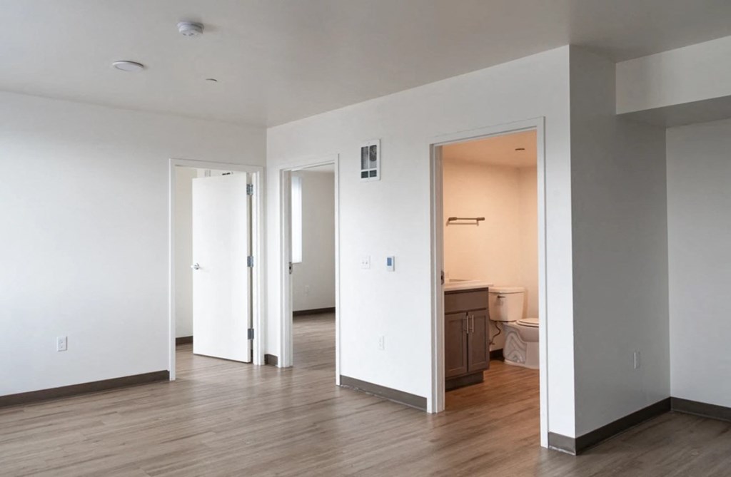 A room with white walls and wooden floors, with a doorway leading to another room.