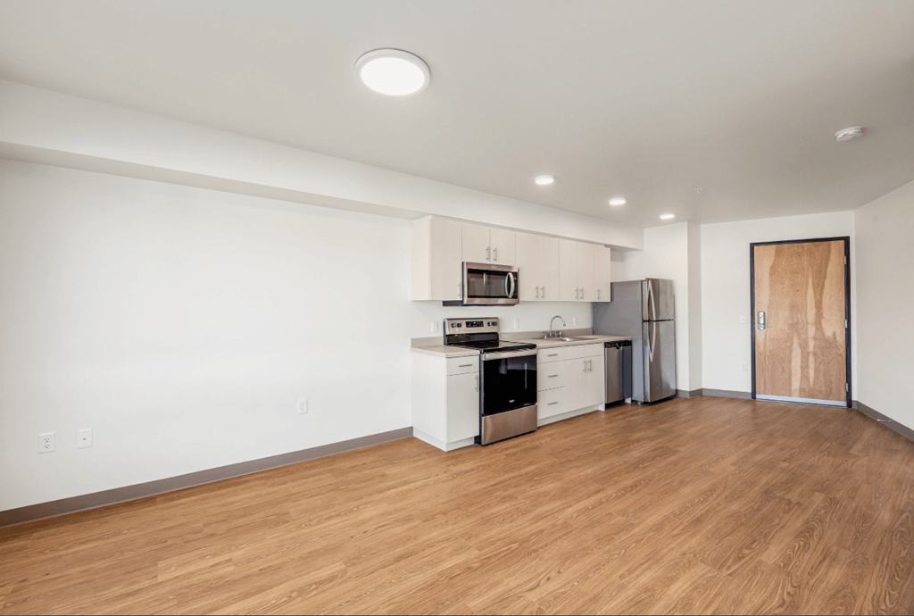 the living room and kitchen of an apartment with wood flooring and white walls and