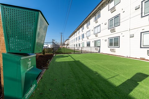 A green trash can is in the foreground of a grassy area in front of apartment buildings.