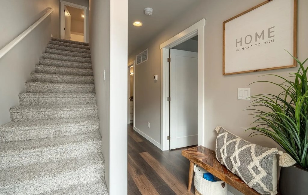 A staircase with a carpeted runner leads to a landing with a wooden bench and a framed sign that says "HOME is next to you.".