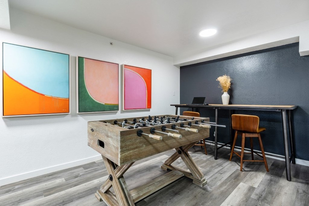 A room with a foosball table, a desk, and three colorful paintings on the wall.
