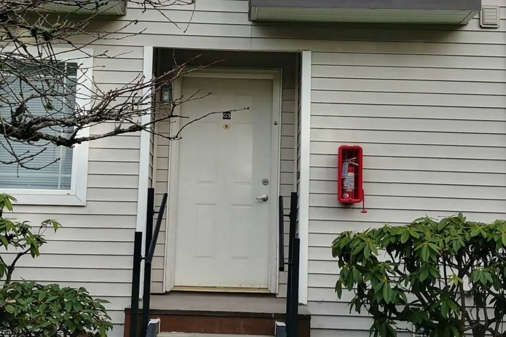 A white door is on the right side of a house with a red fire extinguisher mounted on the wall.