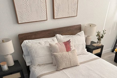 A bed with white pillows and a wooden headboard is in the foreground with two framed artworks on the wall behind it.