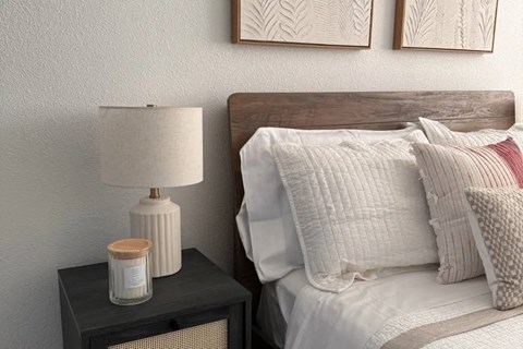 A lamp with a white shade and a beige lampshade cover is on a nightstand next to a bed with white bedding.