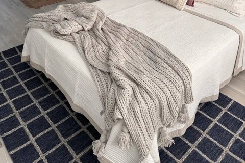 A white bed with a grey knitted blanket on it.