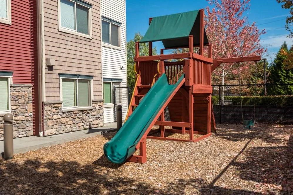 A wooden play structure with a green slide in front of a two-story building.