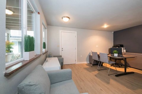 A living room with a grey couch, a table with a laptop, and a window with white blinds.
