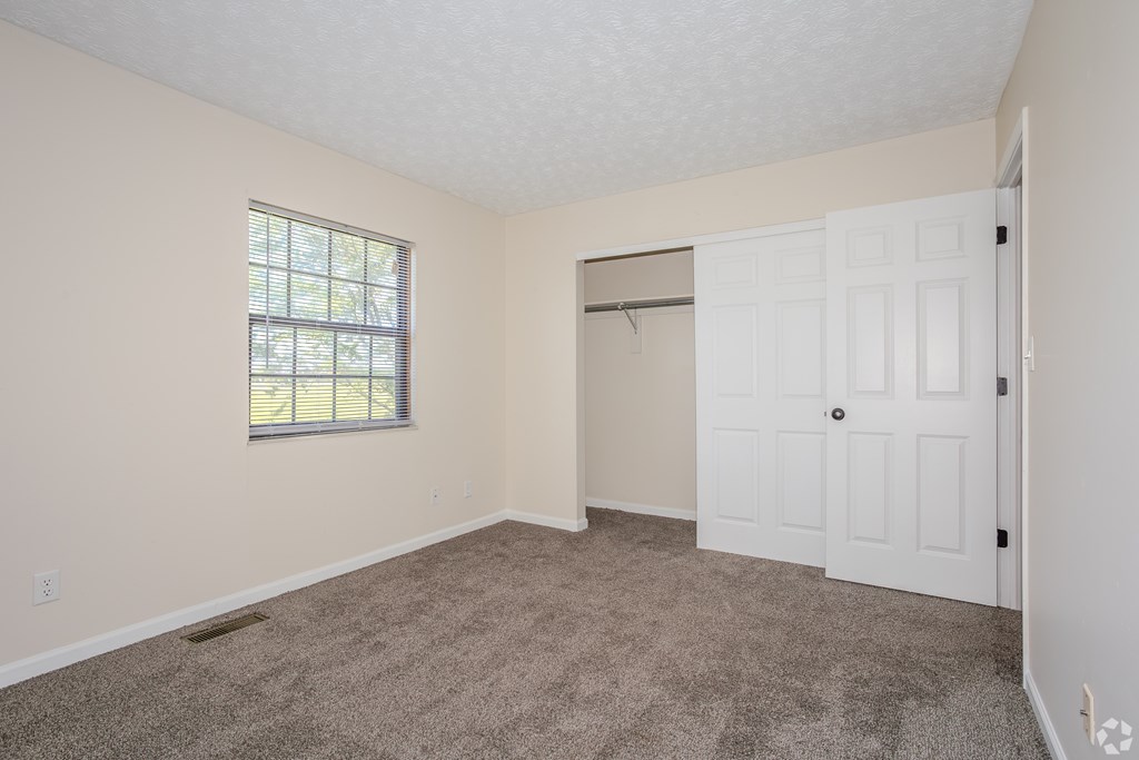 A room with a carpeted floor, a window with blinds, and a white door.
