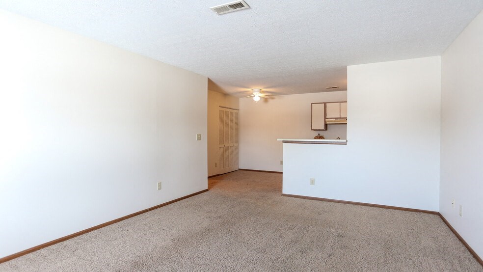 the living room and kitchen of an apartment with white walls and carpet