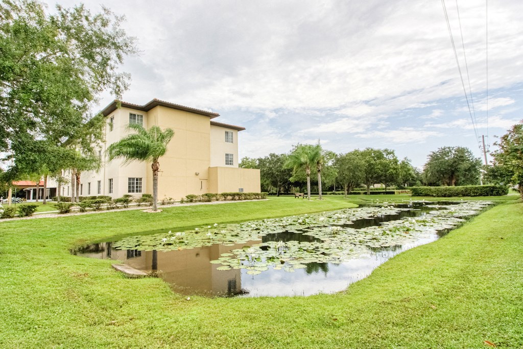pond with lily pads, lush grass, and mature trees beside Holy Cross Manor I