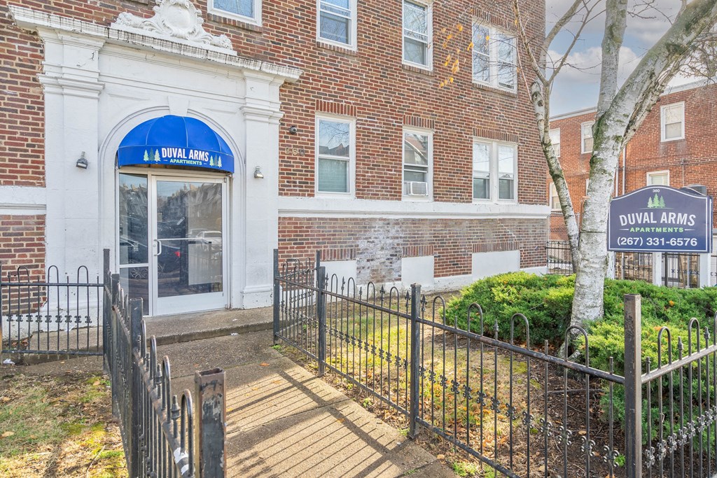 The image shows the entrance to Duval Arms Apartments with a blue signboard and a tree in front.