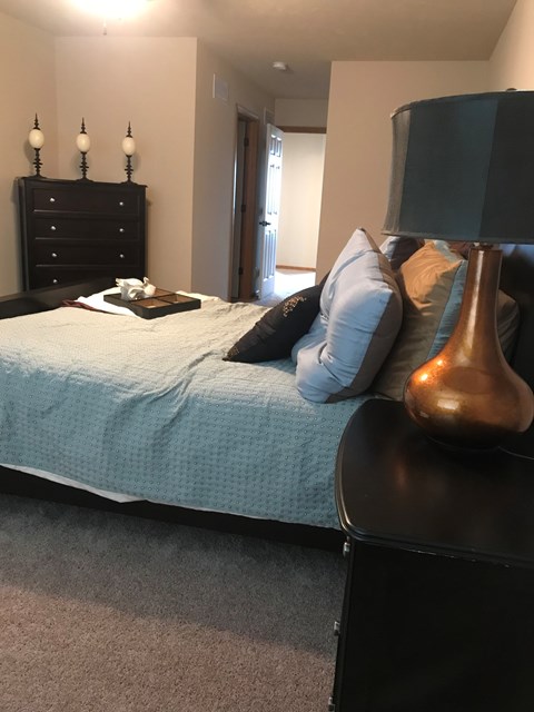 A bedroom with a bed, nightstand, lamp, and pillows.
