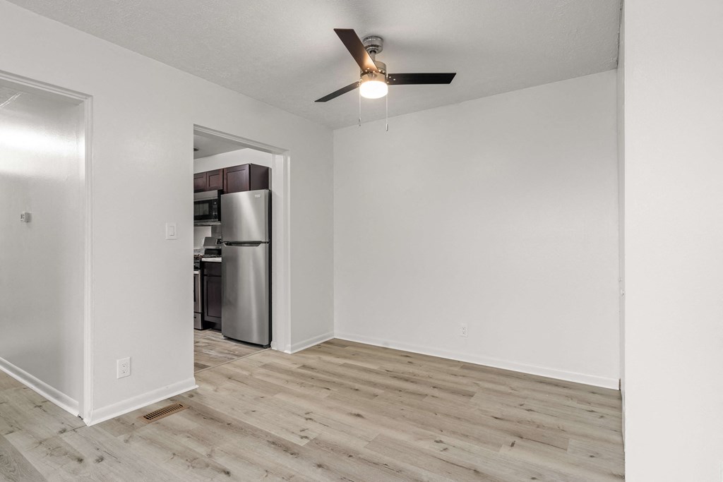 A room with a ceiling fan and a kitchen in the background.