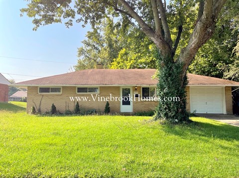 A house with a brown roof and a white garage door is surrounded by green grass and trees.