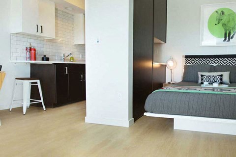 A modern bedroom with a grey bed and a kitchen area with black cabinets.
